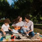 A joyful family enjoying a summer picnic with a ukulele in a sunny park setting.