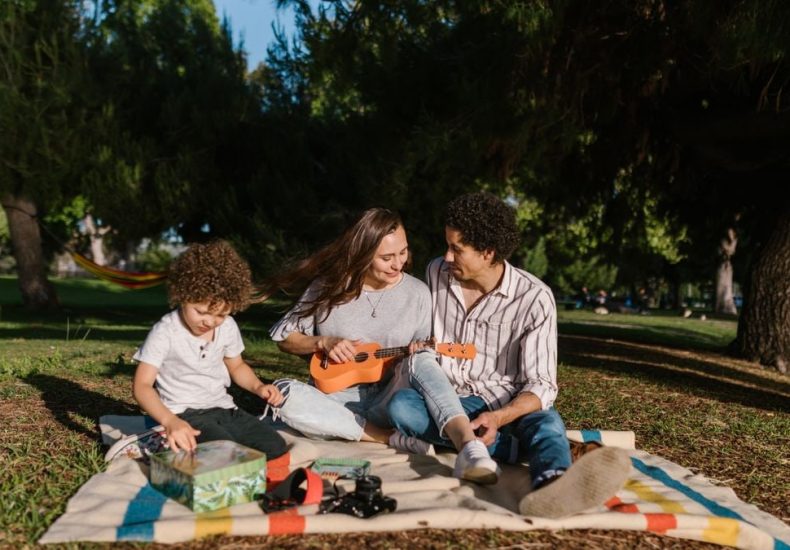 A joyful family enjoying a summer picnic with a ukulele in a sunny park setting.