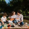 A joyful family enjoying a summer picnic with a ukulele in a sunny park setting.